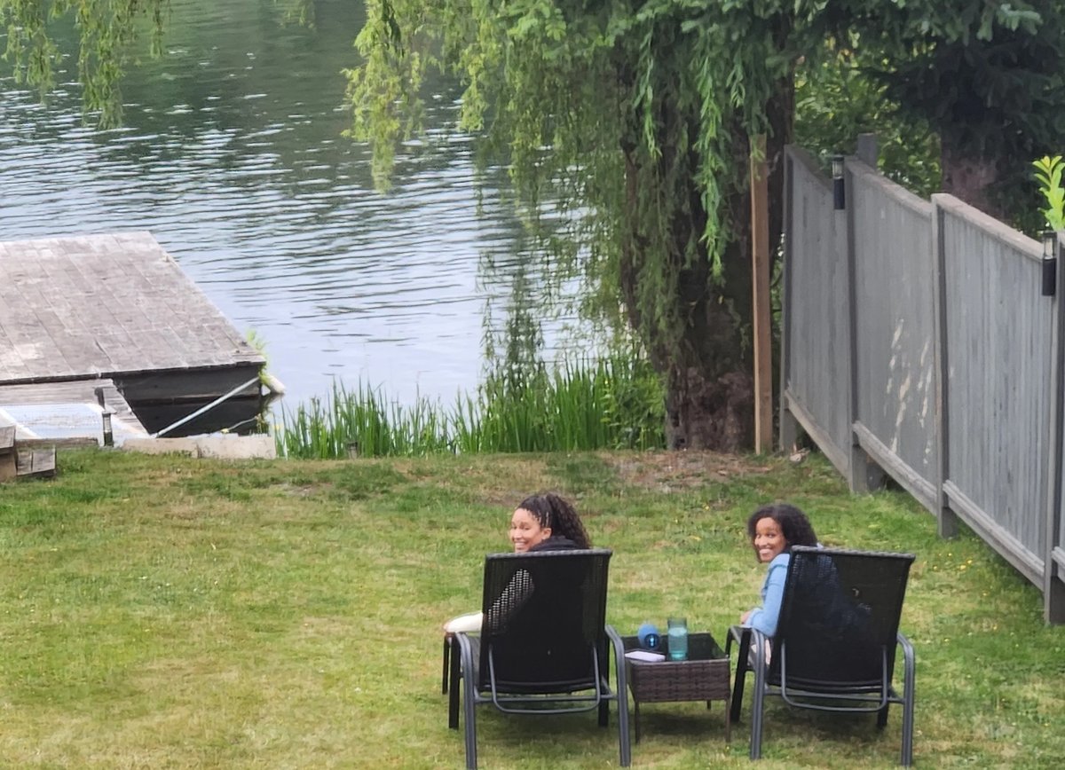 Two people sitting in black lawn chairs on grass facing a waterside backyard with a gray fence and dock visible