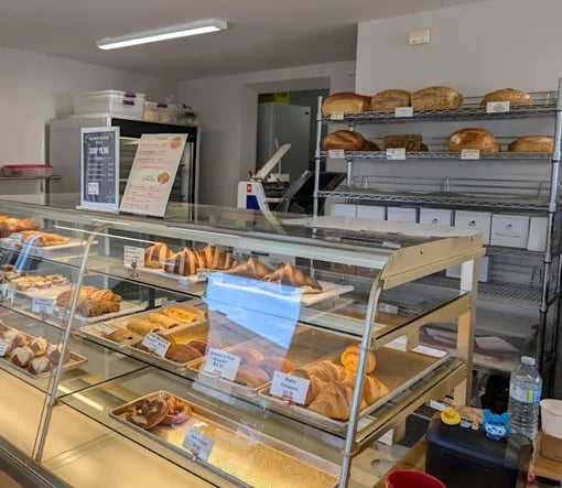 A bakery display case filled with pastries and baked goods, with shelving behind containing bread loaves and rolls