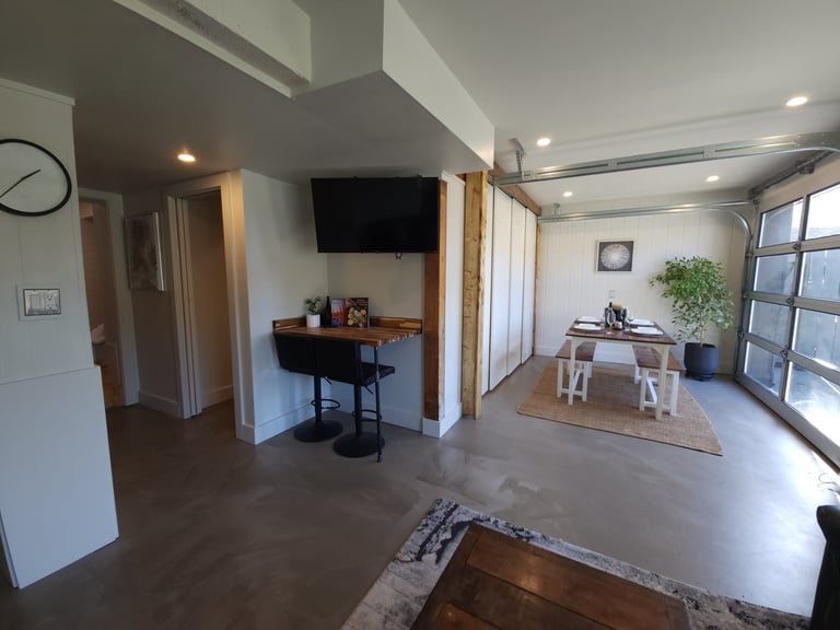 Modern open-concept apartment interior with wall-mounted TV, desk area, dining table, large windows, and polished concrete flooring