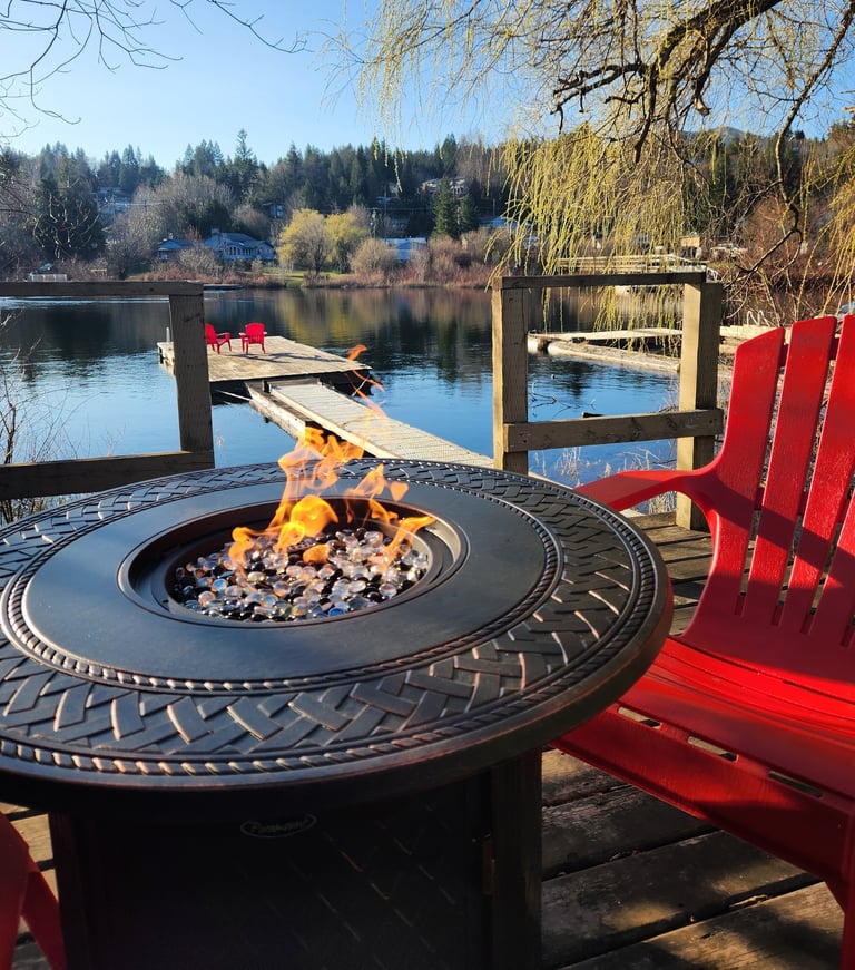 Waterfront fire pit with bright red Adirondack chairs, burning flames, and calm lake with docks and evergreen trees in background
