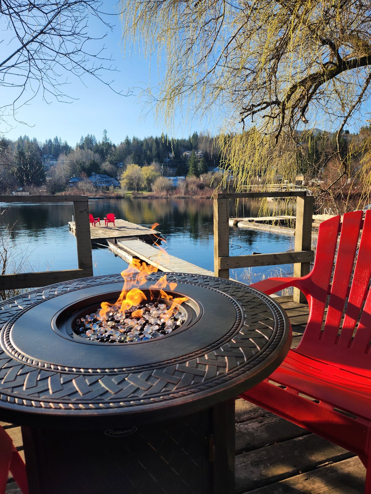 Red Adirondack chairs facing a lit fire pit on a lakeside dock with boats and evergreen trees in background